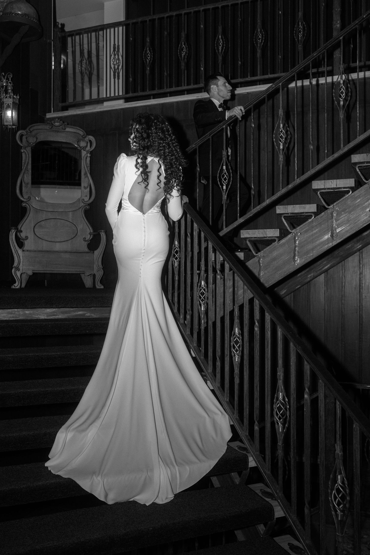 Black and white photo of bride in backless mermaid gown ascending grand staircase with groom.