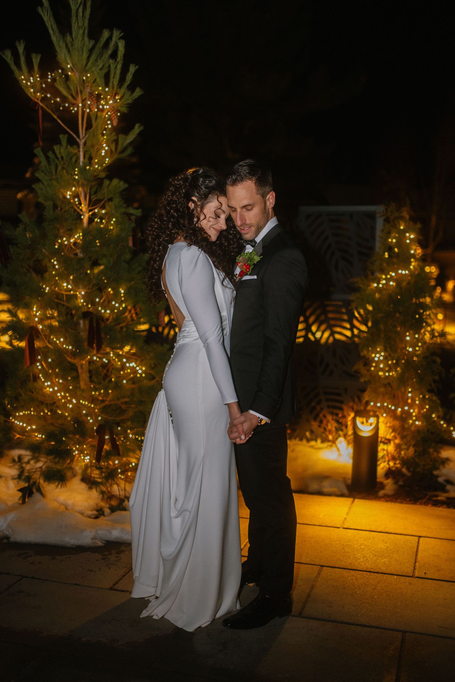 Bride and groom share romantic moment outdoors at night beside Christmas trees adorned with warm golden lights in winter setting.