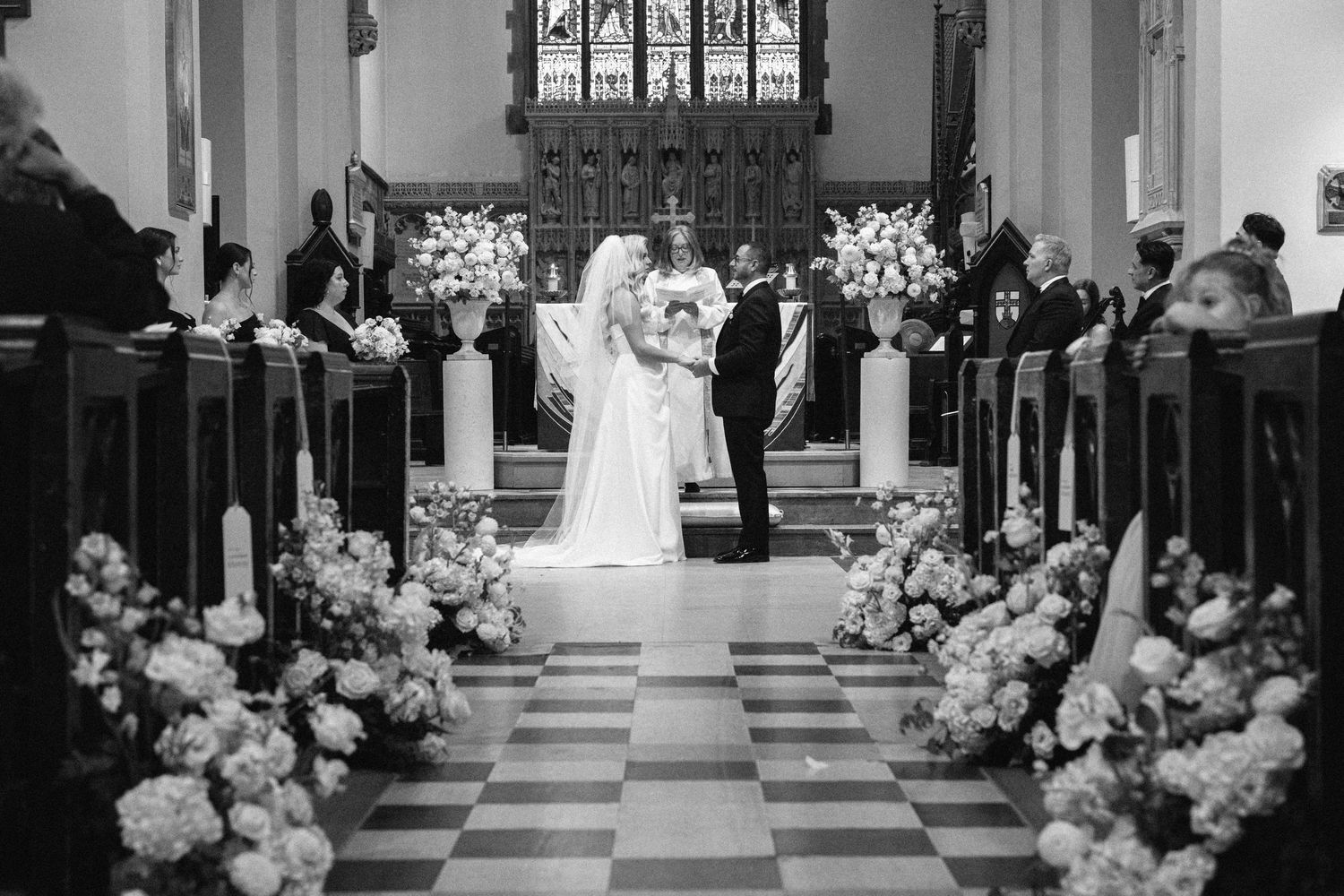 Black and white wedding ceremony photo of couple at altar surrounded by white floral arrangements in church.