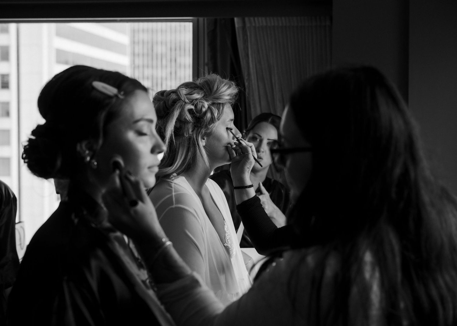 Bridesmaids and bride getting hair and makeup done by stylists in front of a bright window.