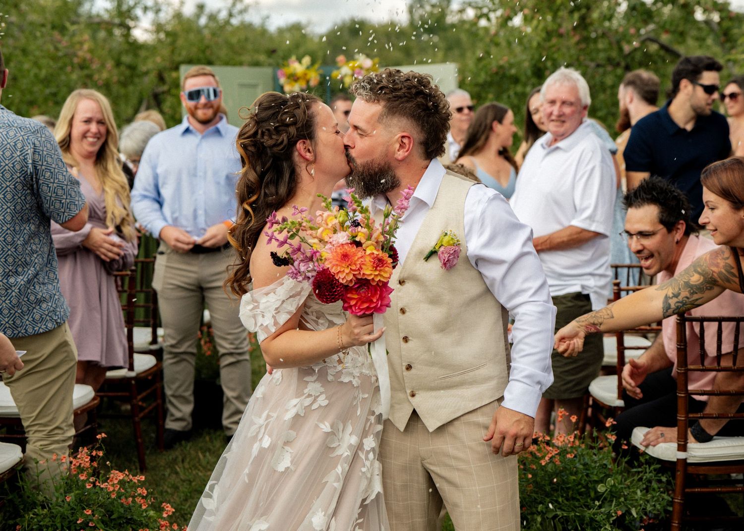Bride and groom share a kiss at outdoor wedding ceremony, bride holding colorful bouquet, guests celebrating in background.