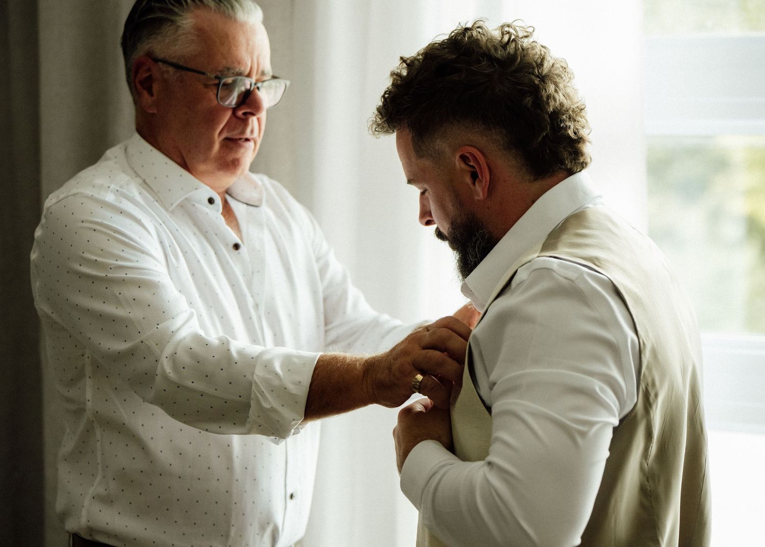 An older man in glasses helps a bearded groom adjust his suit jacket before a wedding ceremony.