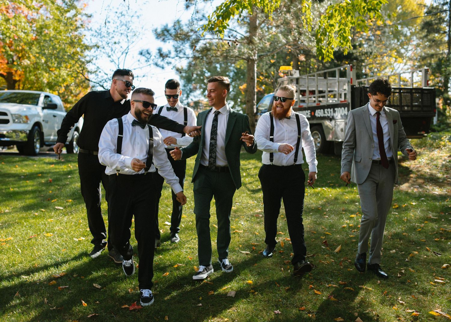 Groomsmen in black suits with suspenders walk outdoors on grass at a wedding celebration.