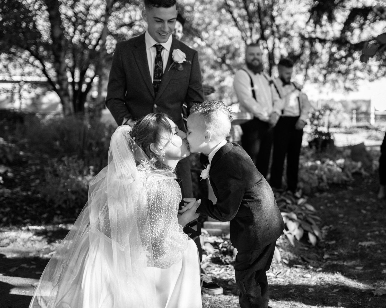 Bride bending down to kiss young child at outdoor wedding ceremony in black and white.