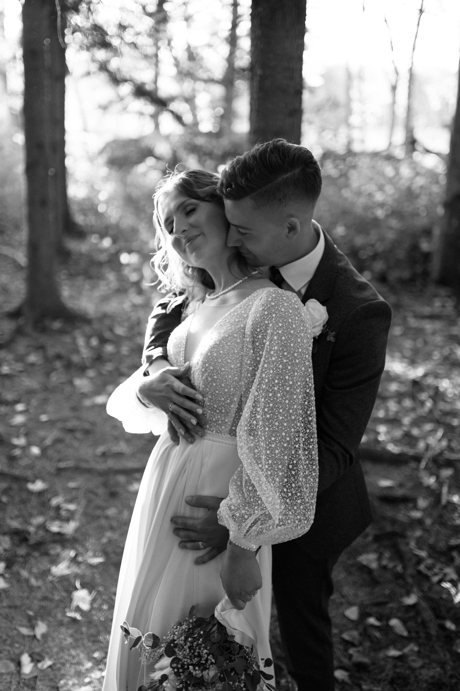Groom embracing bride from behind in a sunlit forest in a black and white wedding photo.
