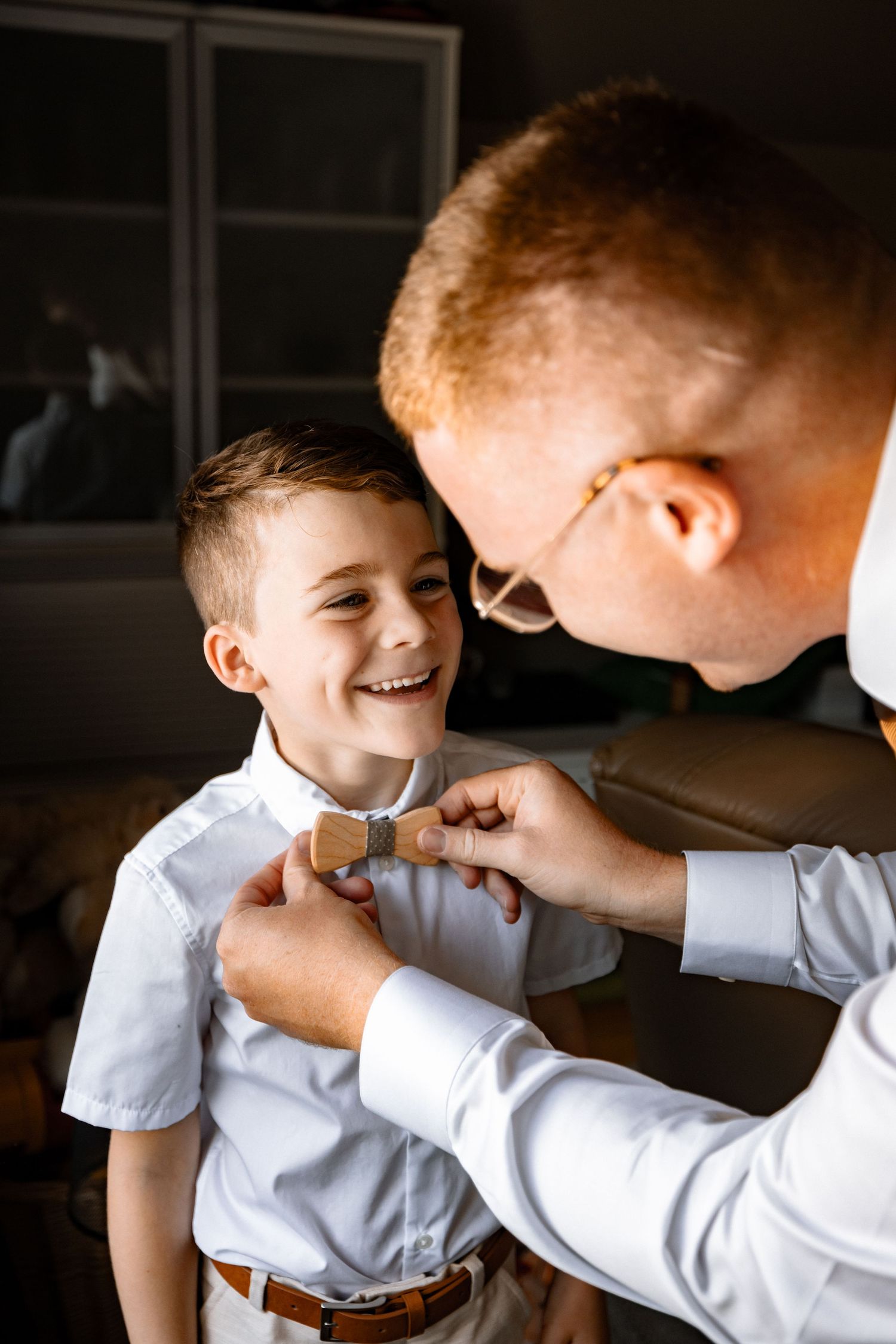 A father adjusts his smiling young son's bow tie, both dressed in white shirts for a formal occasion.