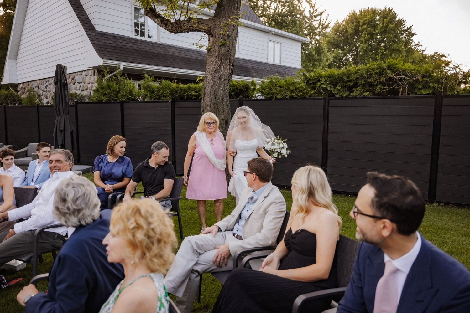 Bride walks down aisle in backyard wedding ceremony with seated guests on sunny day.