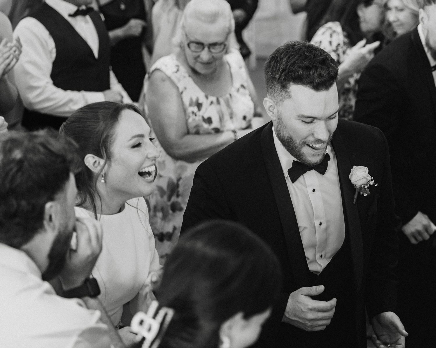 Black and white photo of a laughing bride and groom in a tuxedo celebrating at their wedding reception.