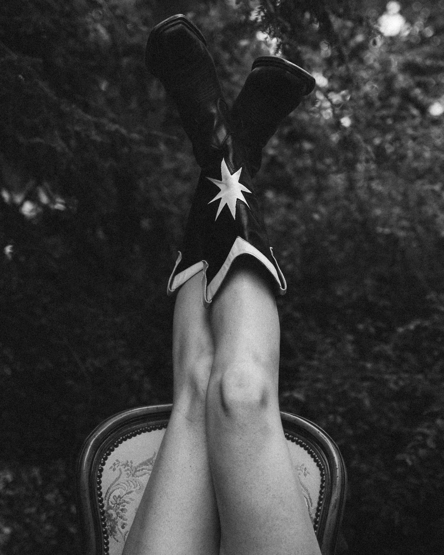 Black and white photo of legs raised up wearing black cowboy boots with white star detail, seated on ornate chair outdoors.