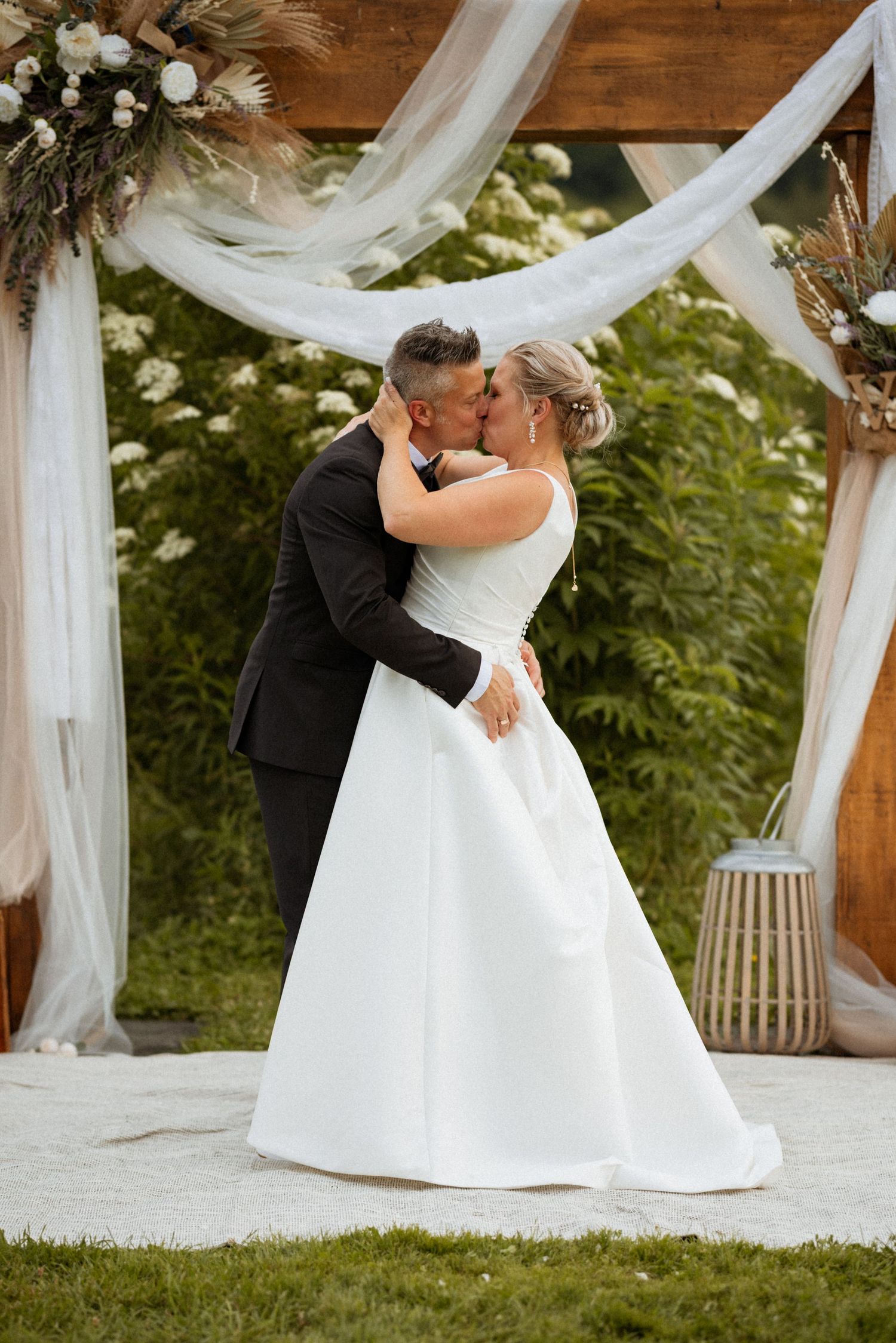 Bride and groom share first kiss under rustic wooden arch with white draping and floral arrangements at outdoor wedding ceremony.