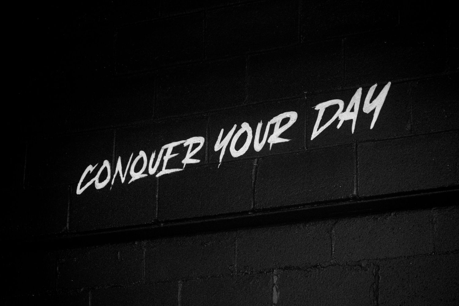 Motivational graffiti-style neon text reading Conquer Your Day on dark brick wall in black and white.