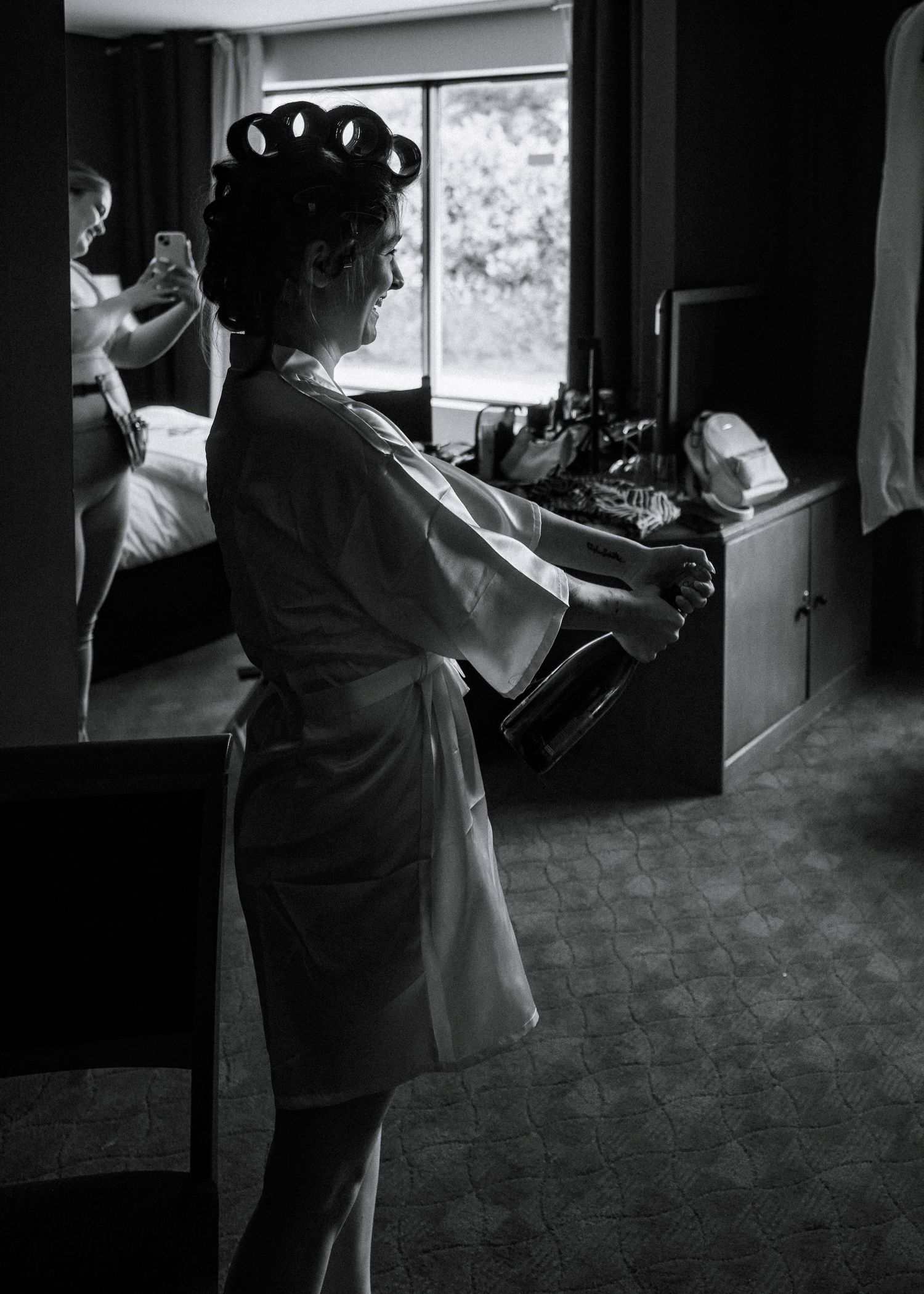 Bride in white robe and hair rollers opening champagne bottle in hotel room before wedding ceremony.