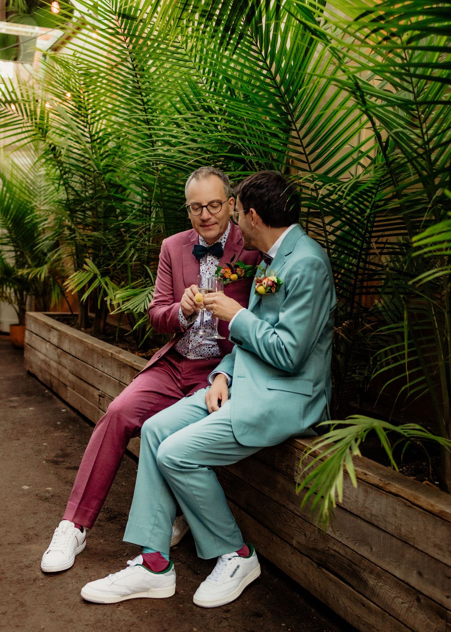 Two grooms in pink and mint suits toasting with champagne while seated on a bench among lush palms.