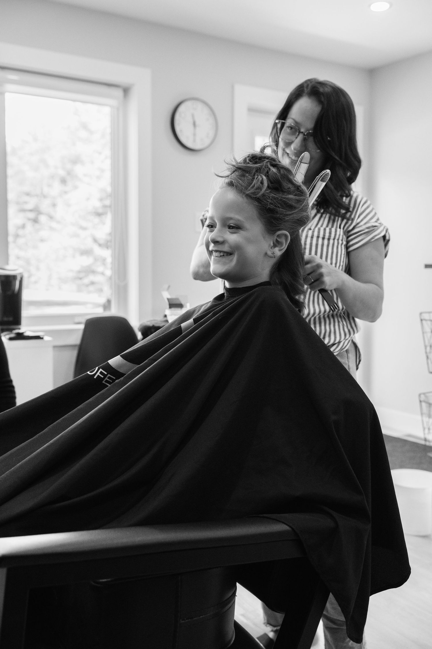 A smiling child getting a haircut at a salon, draped in a black cape, while a hairstylist combs their hair.