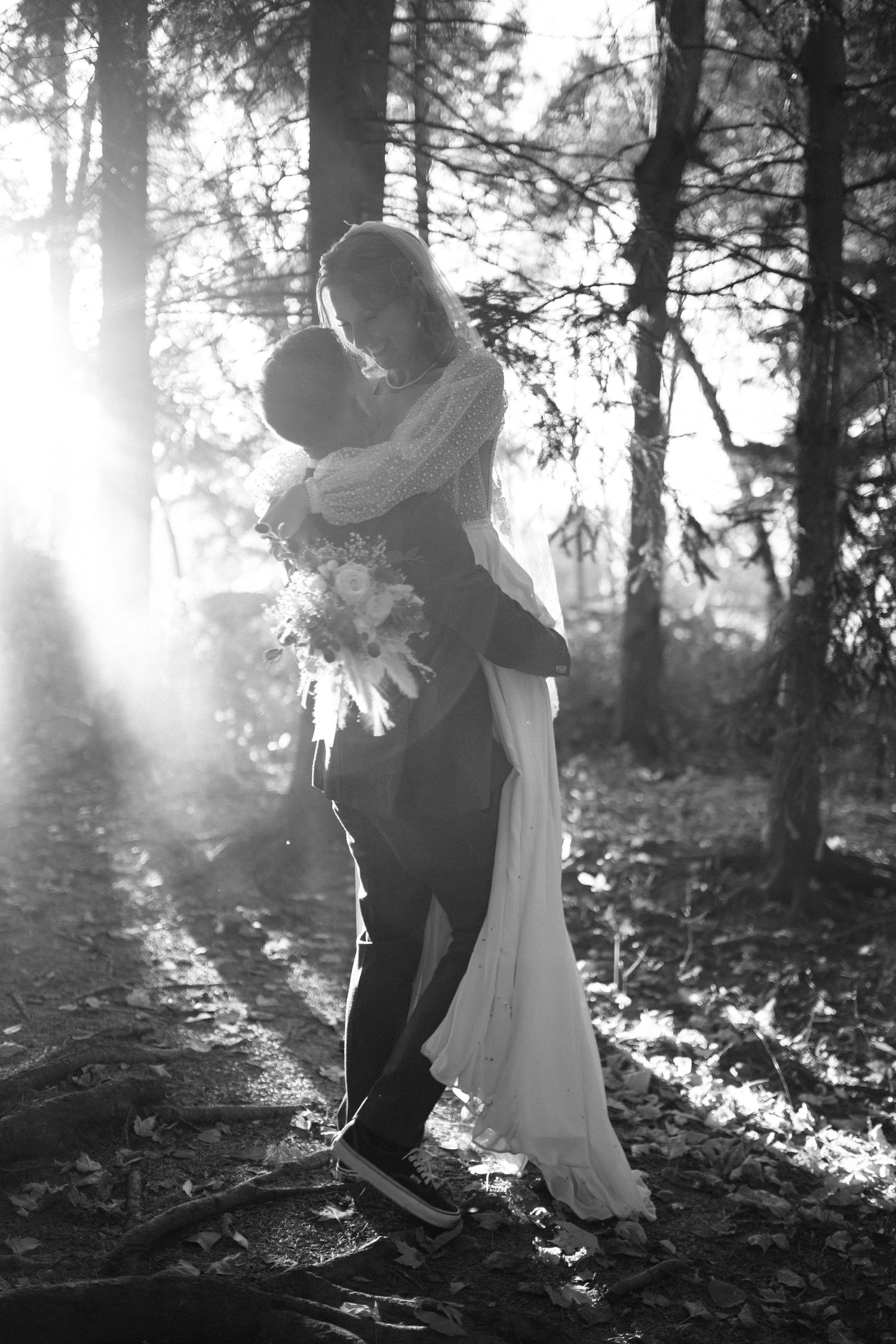 Black and white photo of bride and groom embracing in sunlit forest with wedding bouquet.