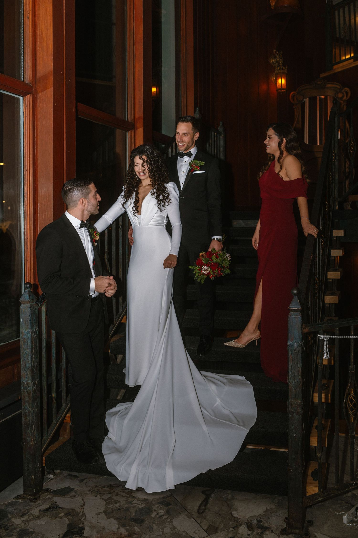 Bride in white gown and groom in tuxedo pose on steps with wedding party guests at elegant evening venue.
