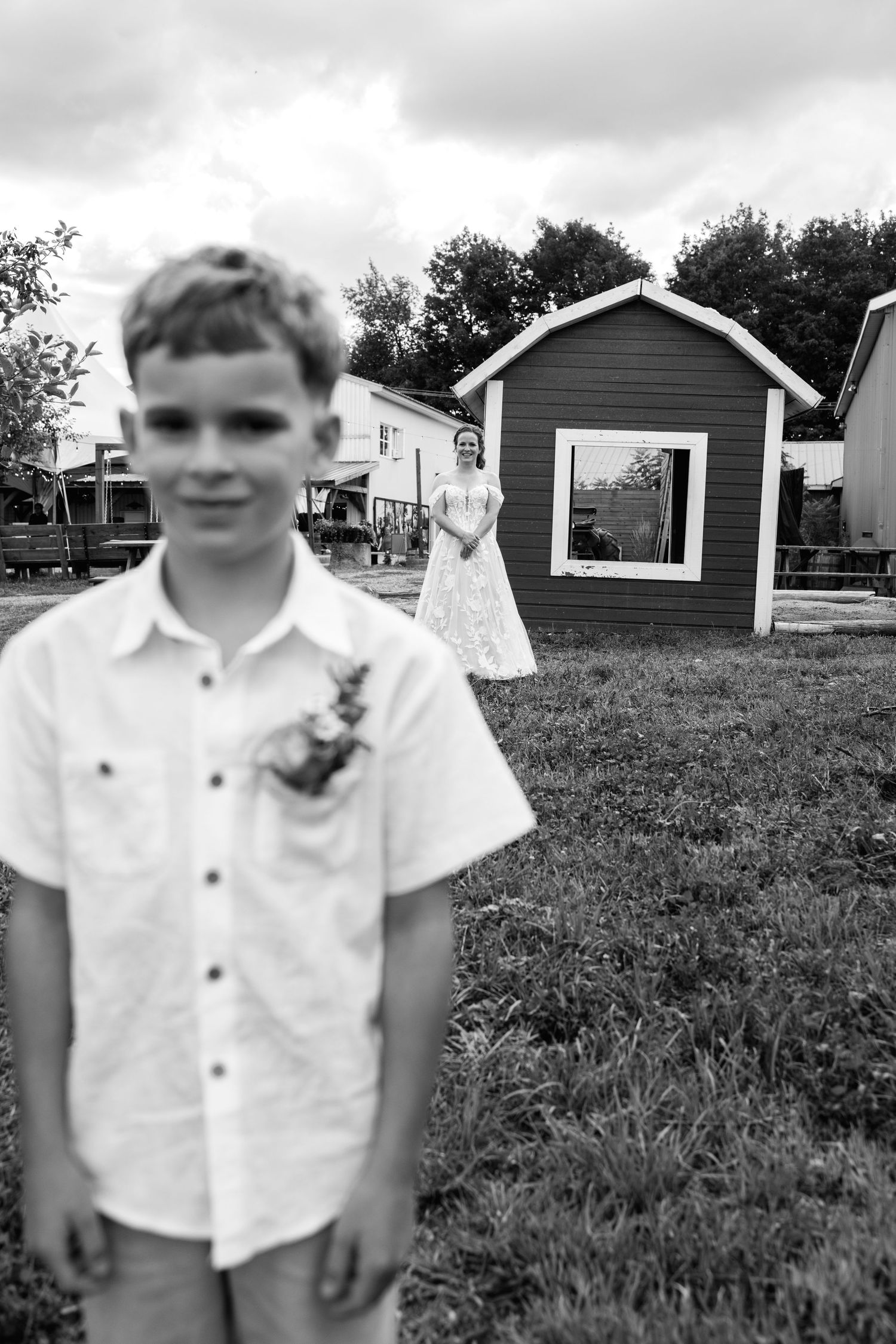 A young boy in a white shirt with a boutonniere stands in focus at a wedding, with a bride posing near a small shed behind him.