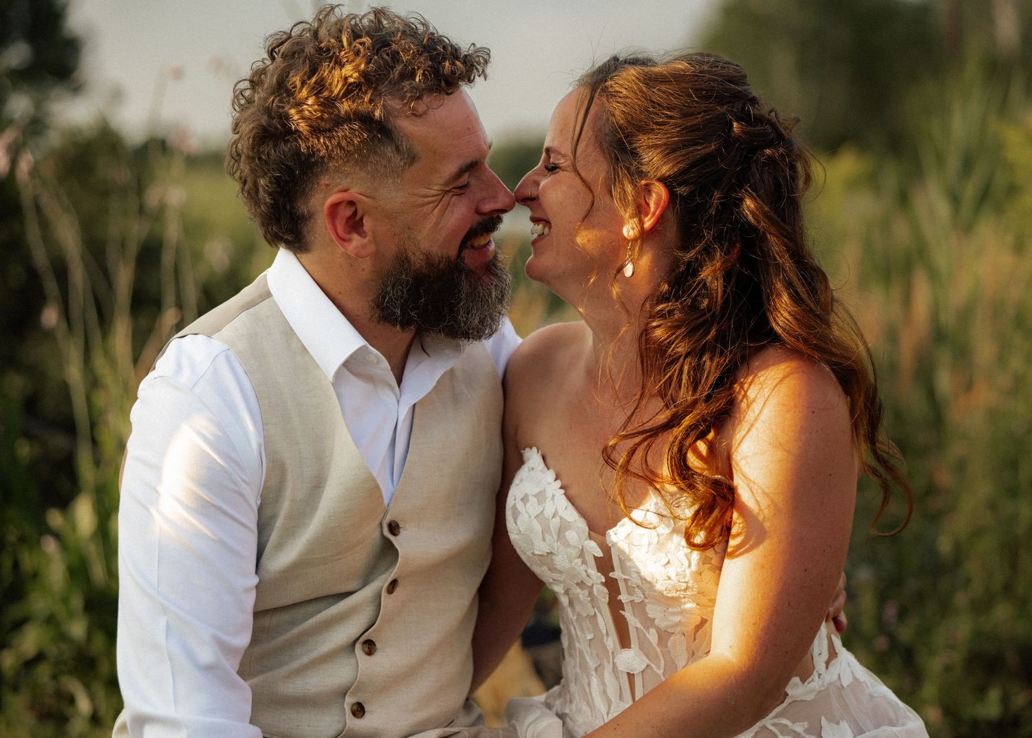 Smiling bride and bearded groom share a romantic nose-to-nose moment in golden outdoor light.