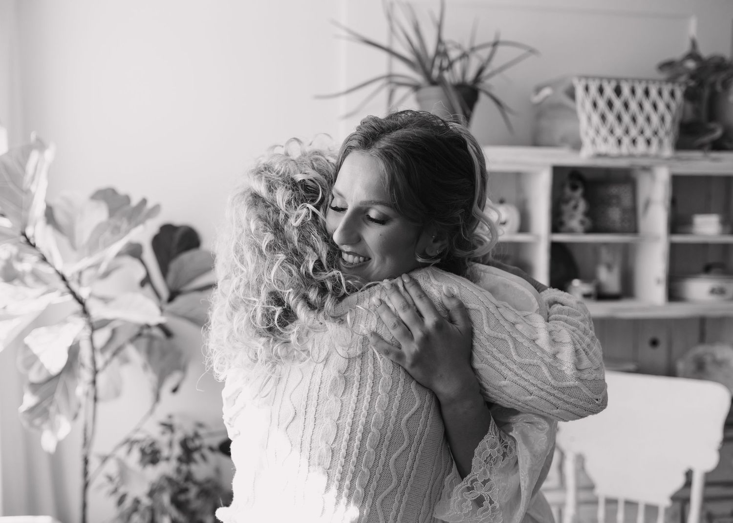 Two women share a warm embrace, smiling joyfully in a bright room with plants and shelving in black and white.