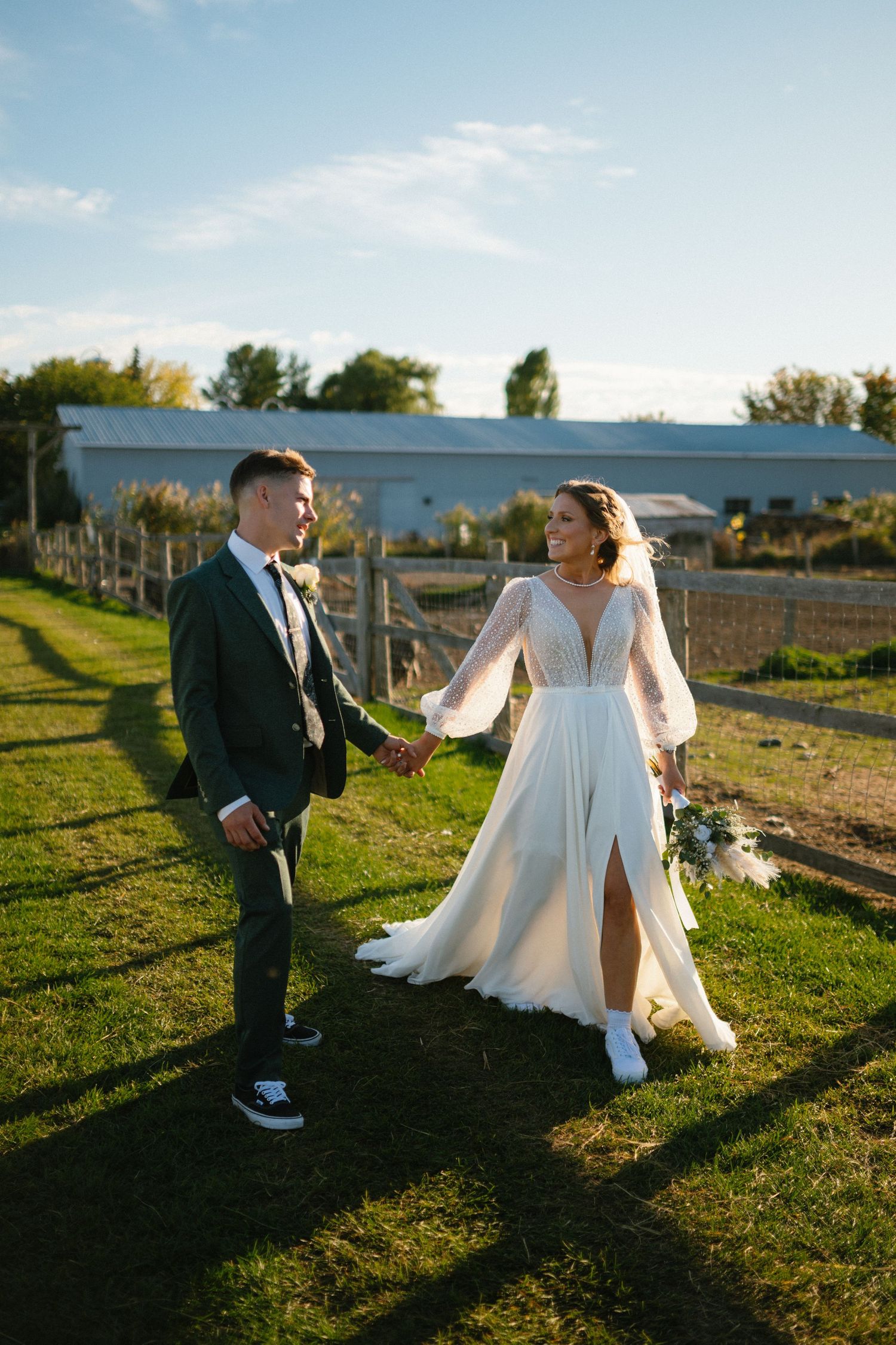 A bride and groom hold hands walking through a sunny farm venue, she in a white long-sleeve gown, he in a dark green suit.