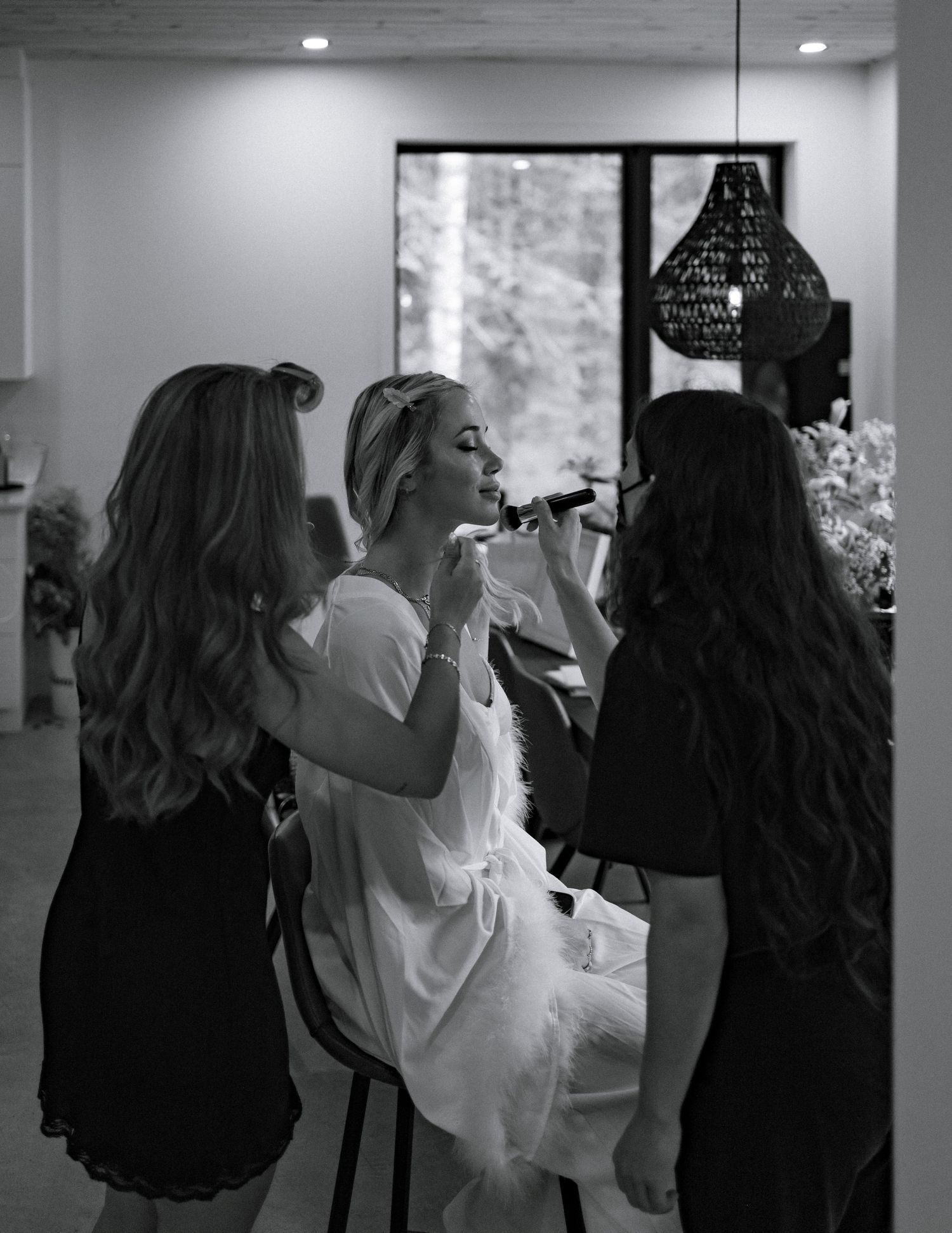 Black and white photo of a bride getting makeup applied by two stylists before her wedding ceremony.