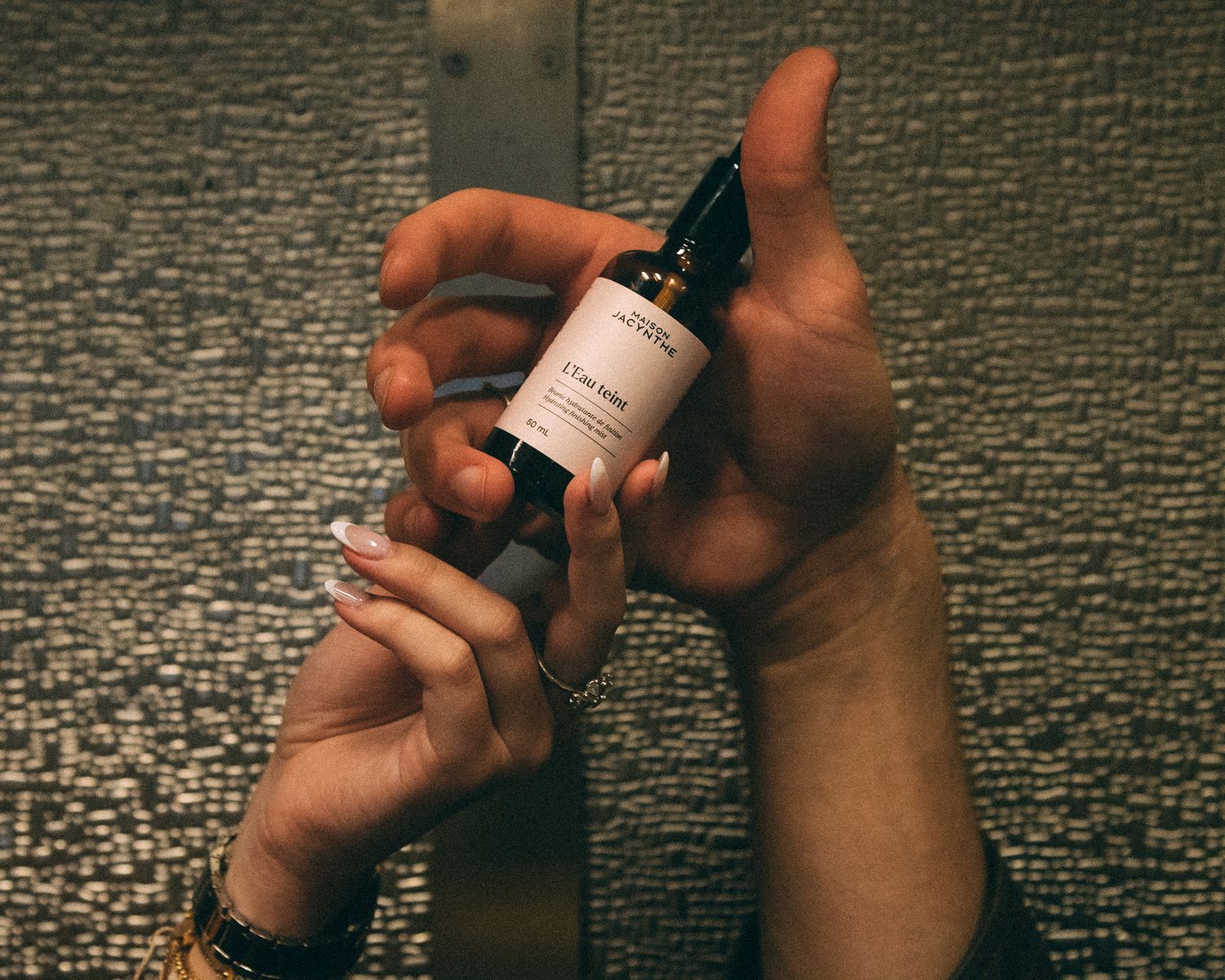 Hands holding a small skincare serum bottle against a textured mosaic tile background.