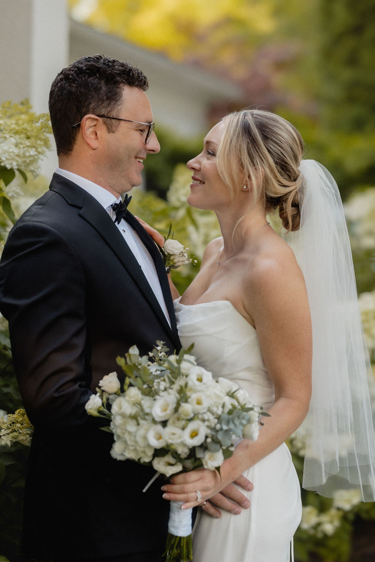 Smiling bride and groom share a romantic moment outdoors, bride holding white bouquet, groom in black tuxedo.