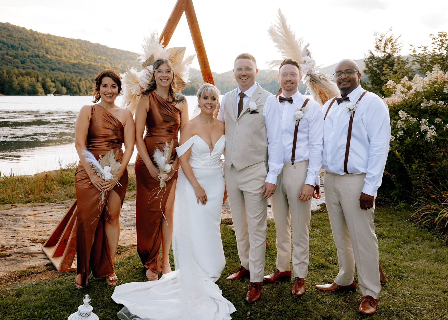 Wedding party poses by boho triangle arch near lake, bridesmaids in rust satin gowns, groomsmen in tan suits.