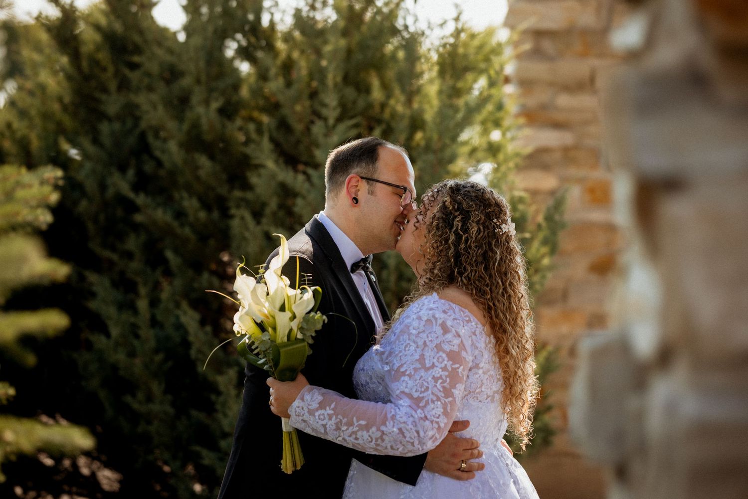 Newlywed couple sharing a kiss outdoors, bride holding white lily bouquet in lace gown.