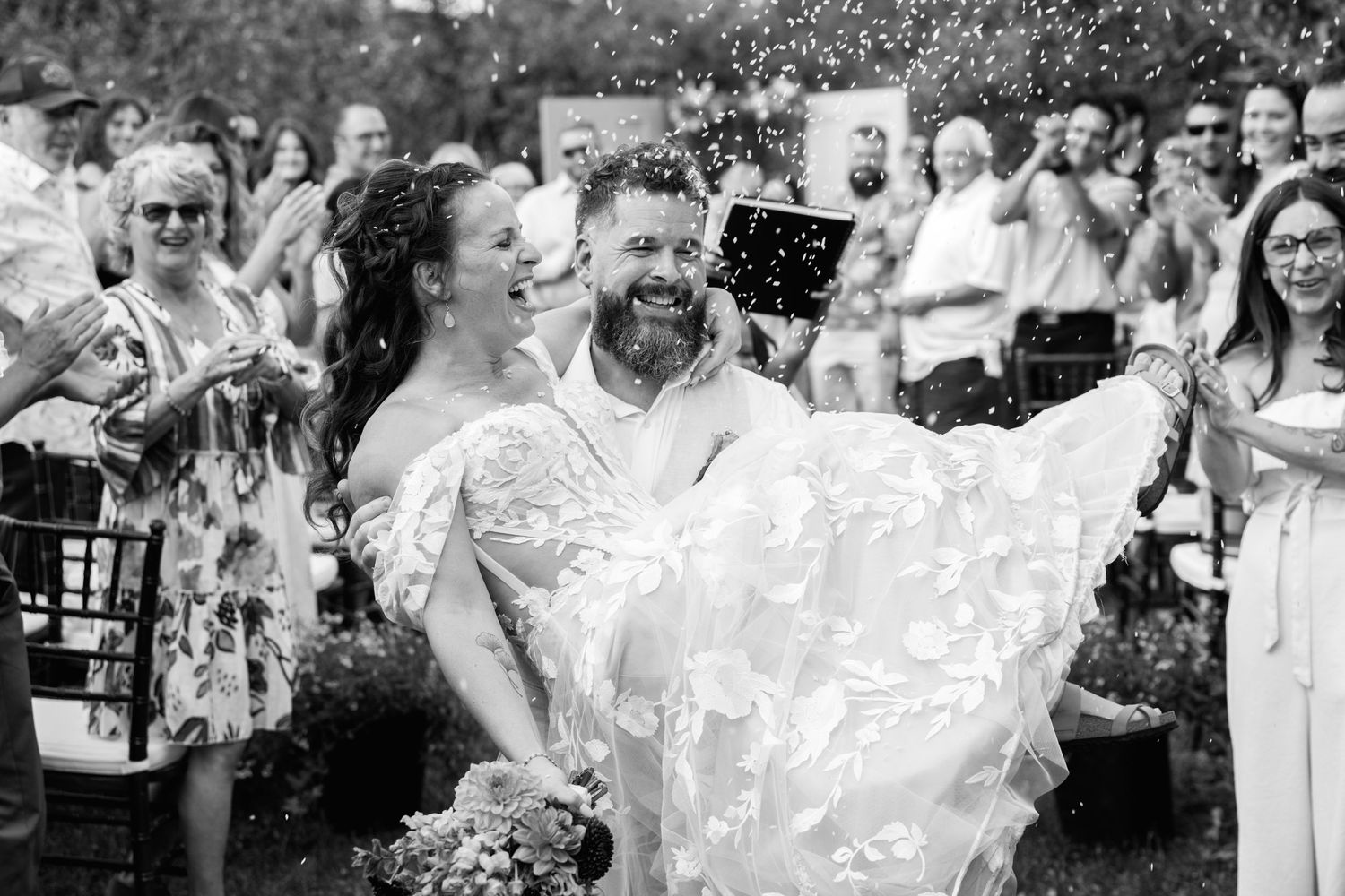 Black and white photo of joyful bride and groom walking through confetti shower at outdoor wedding.