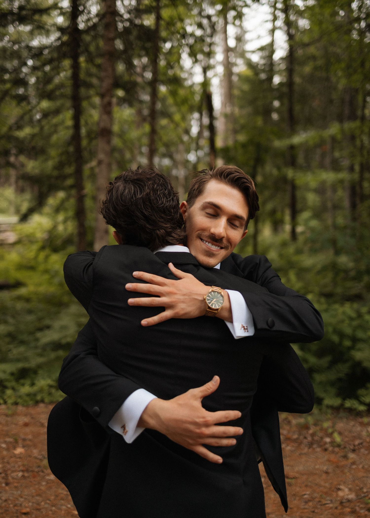 Two men in black suits share a warm hug in a forest setting, one smiling at the camera wearing a wooden watch.