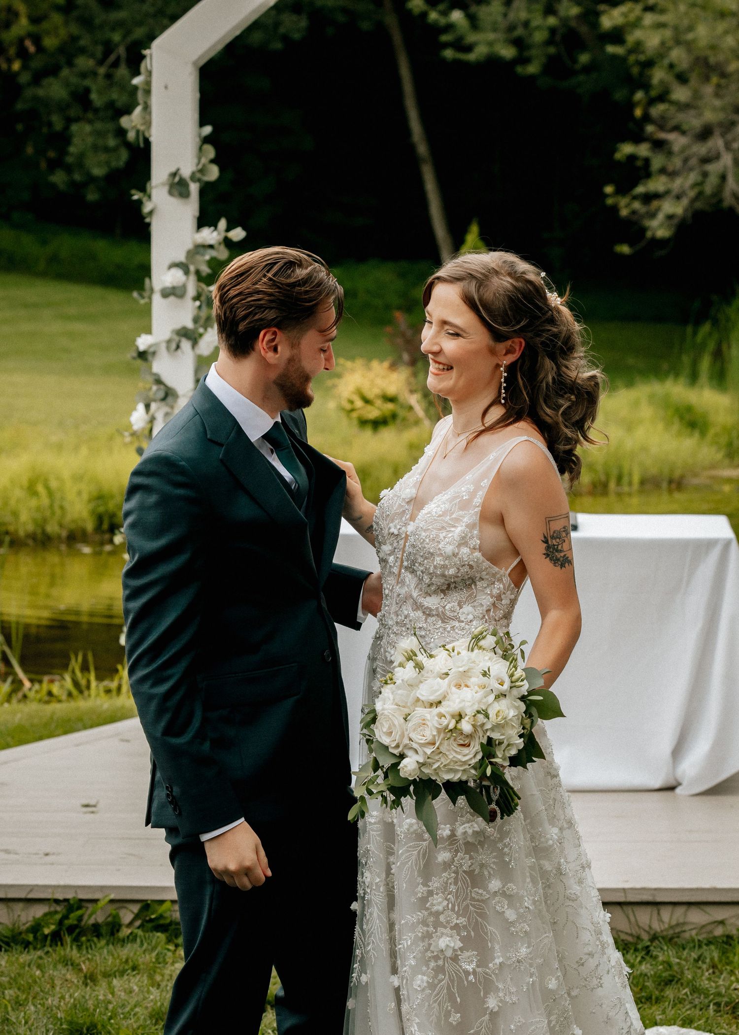 Bride and groom share a joyful moment outdoors, she holds white bouquet in lace gown, he wears navy suit.