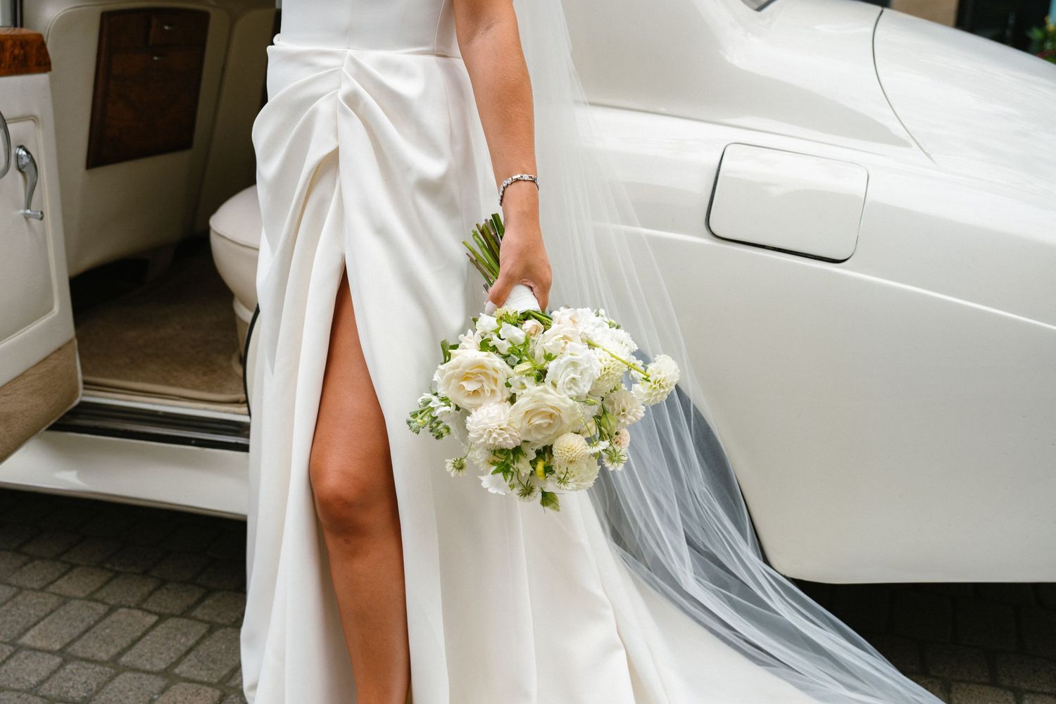 Bride in white satin gown with thigh slit holding elegant white floral bouquet beside a luxury white car.