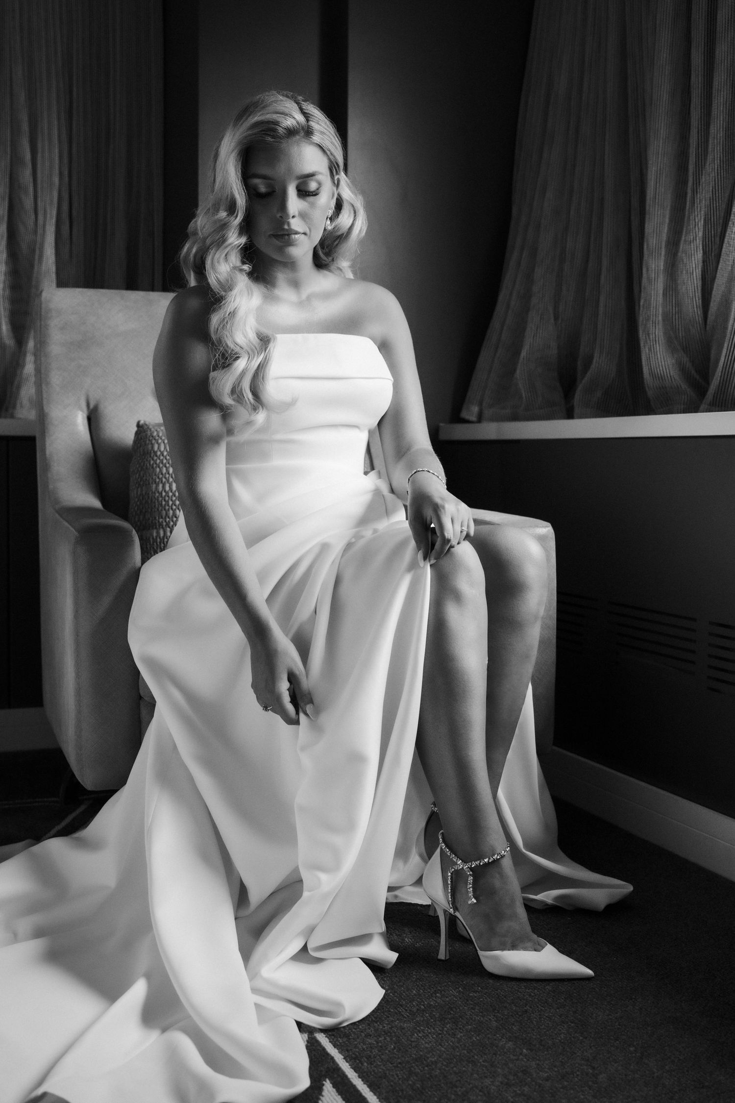 Elegant bride in strapless white gown seated in armchair adjusting her heels, black and white photo.