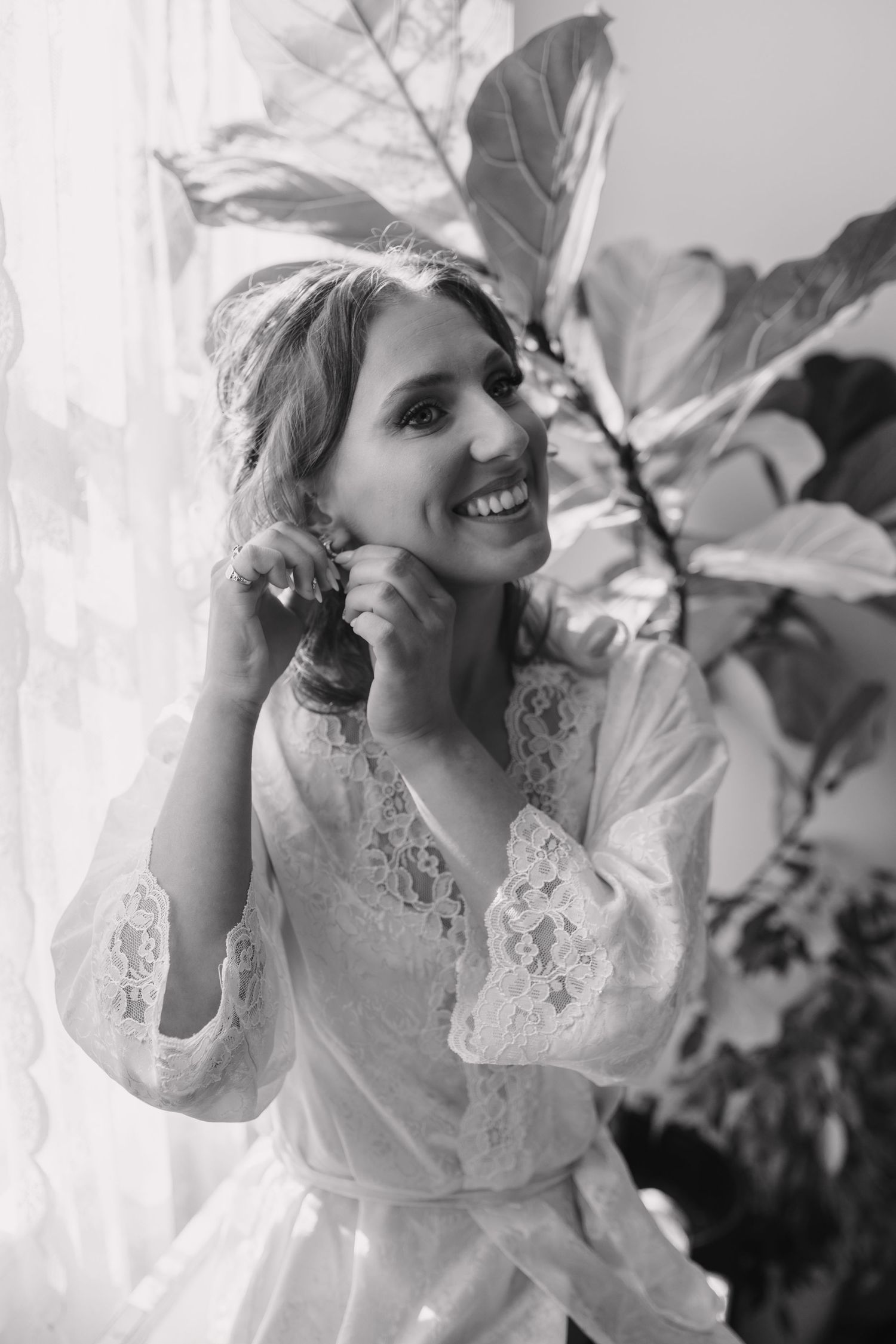 Smiling bride in lace robe adjusting earring by window with tropical plant, black and white.