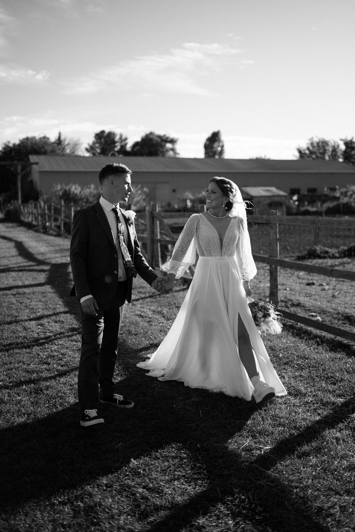 Black and white photo of a bride and groom holding hands at a farm wedding venue, walking together in golden sunlight.