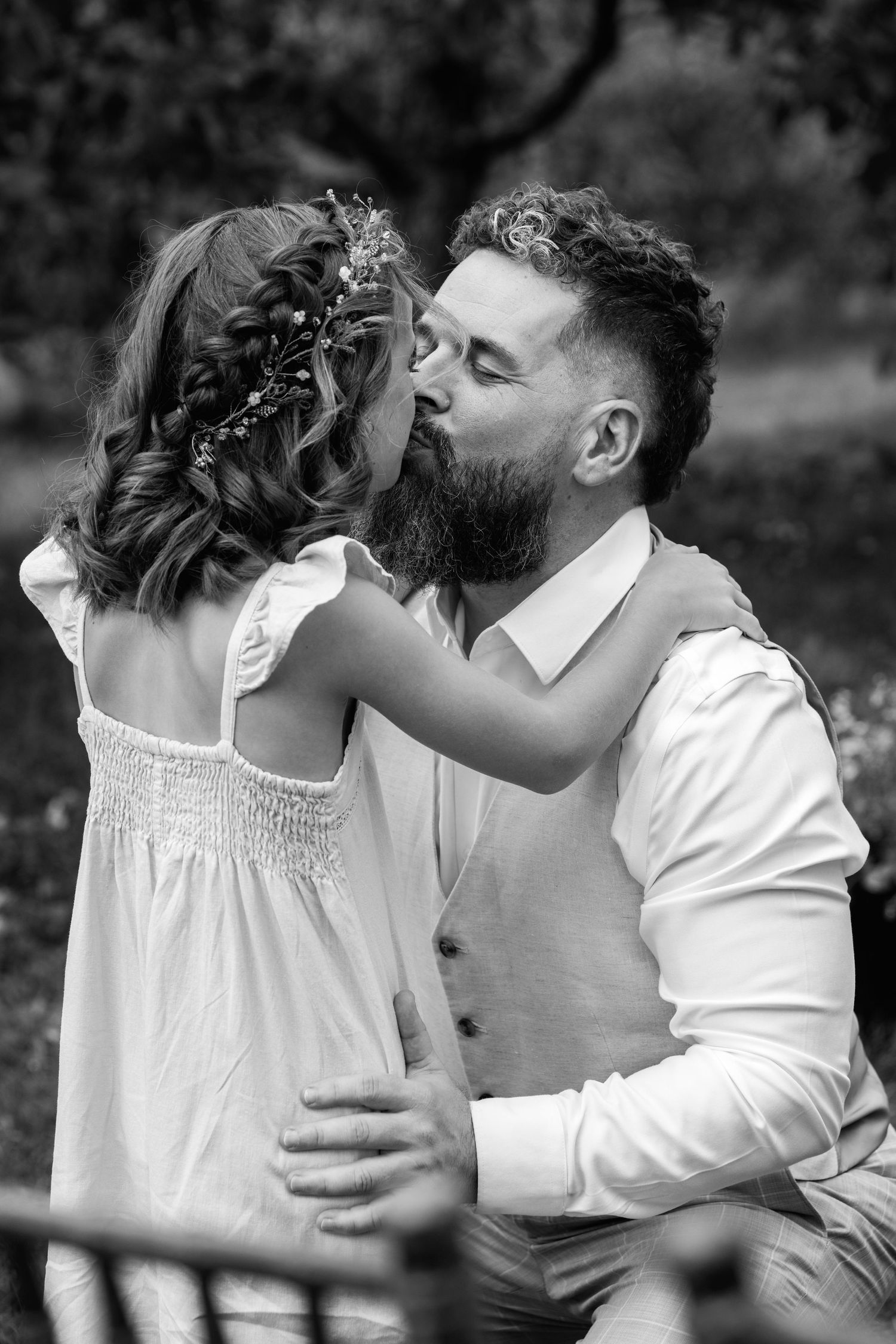 Bearded groom in white suit tenderly kisses a flower girl with braided hair at an outdoor wedding.