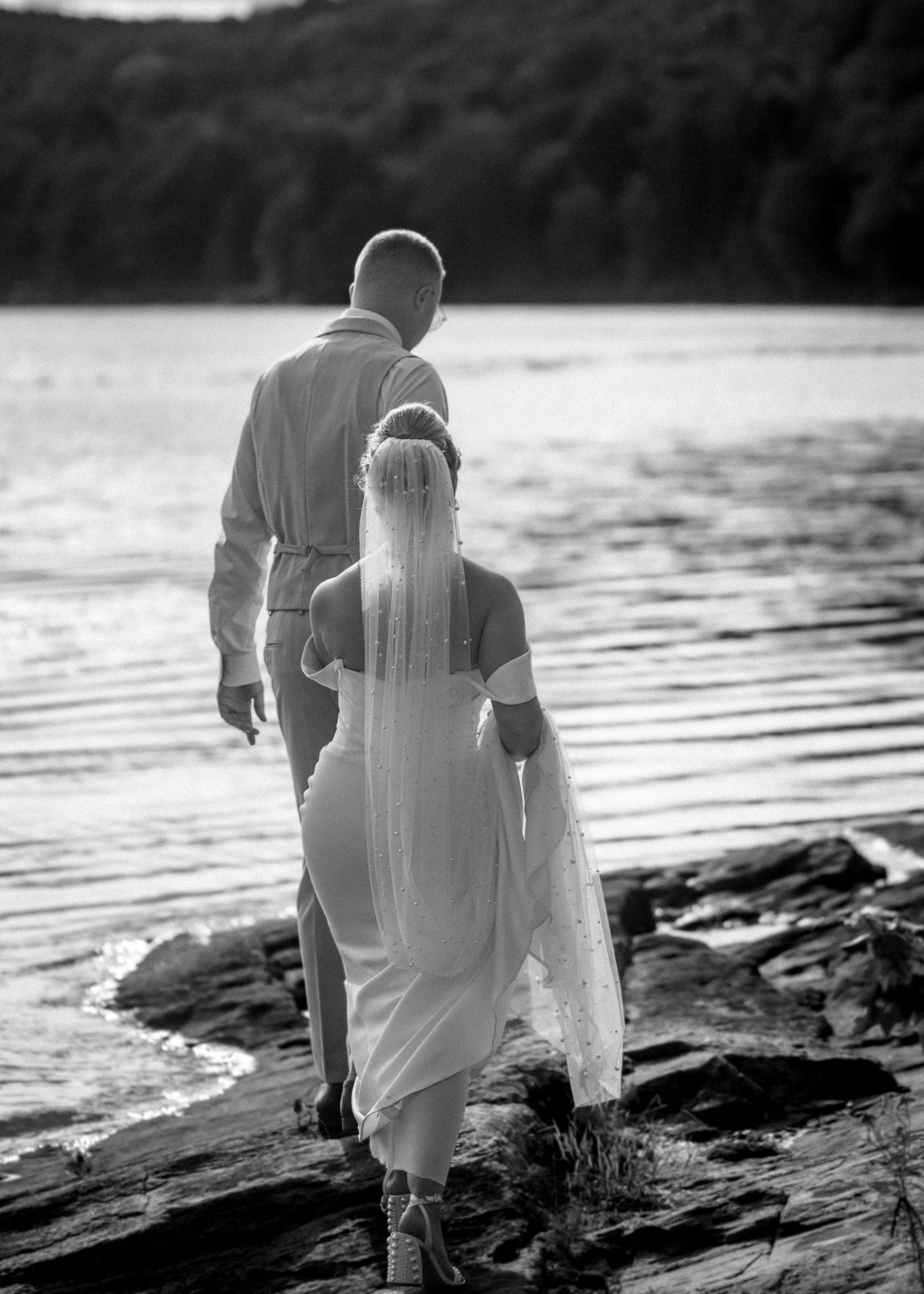 Bride and groom walk along rocky lakeside shore after wedding ceremony, black and white photo.