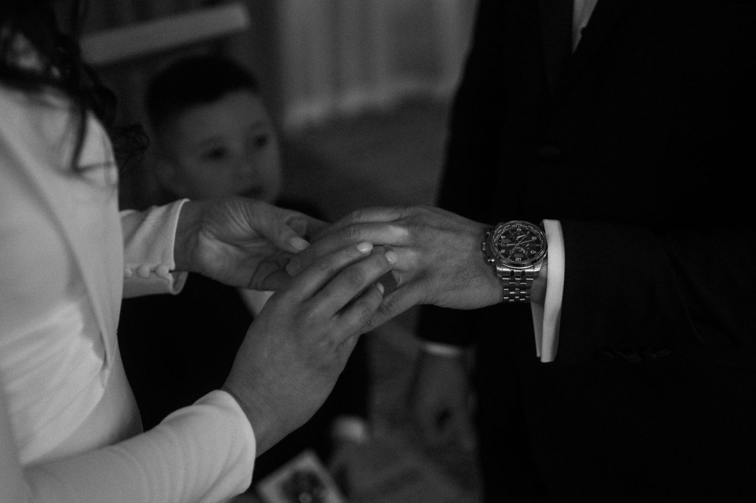 Black and white photo of a couple exchanging wedding rings during a ceremony, with a child visible in the background.