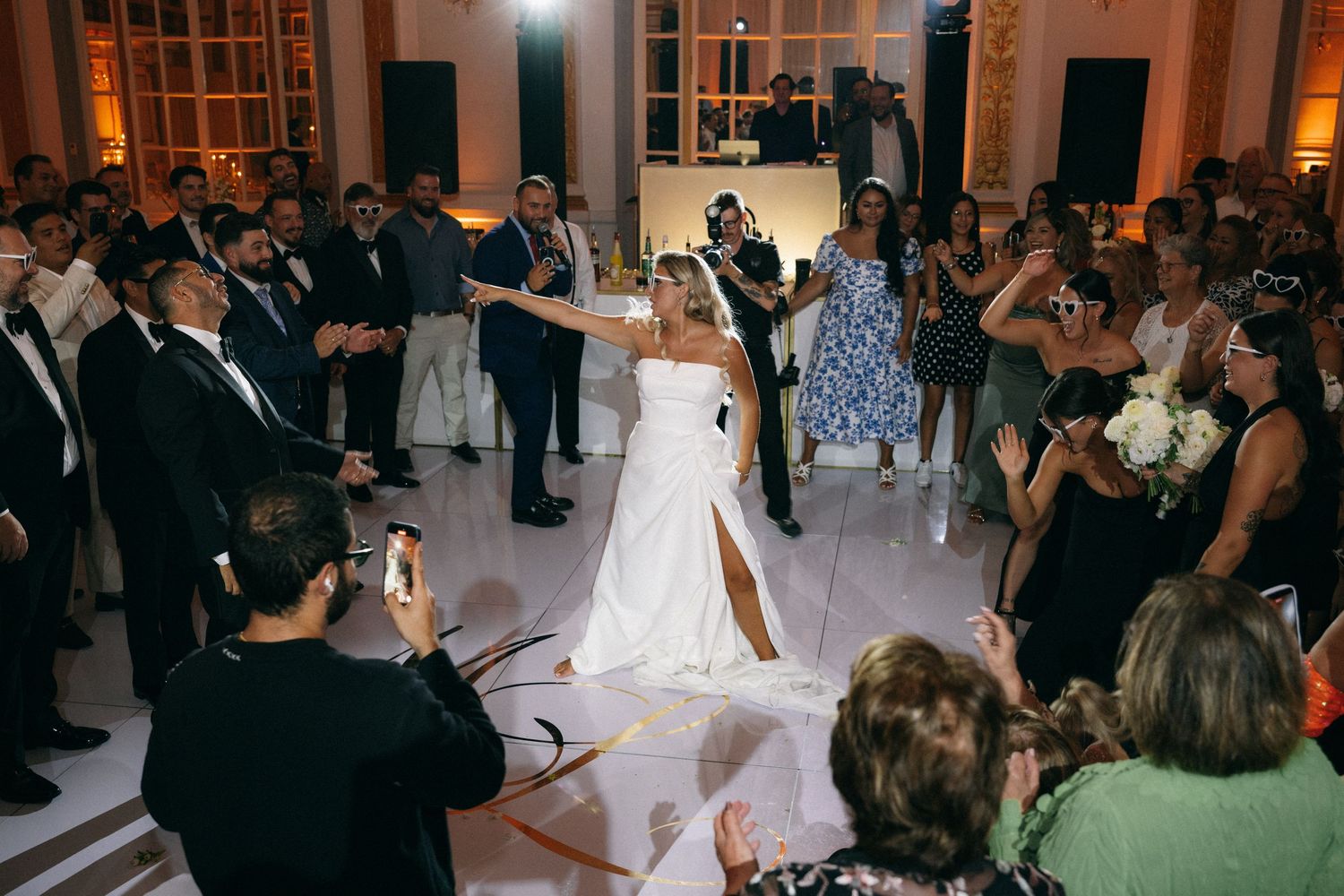 Bride in white strapless gown with slit dances joyfully surrounded by cheering guests at elegant wedding reception.