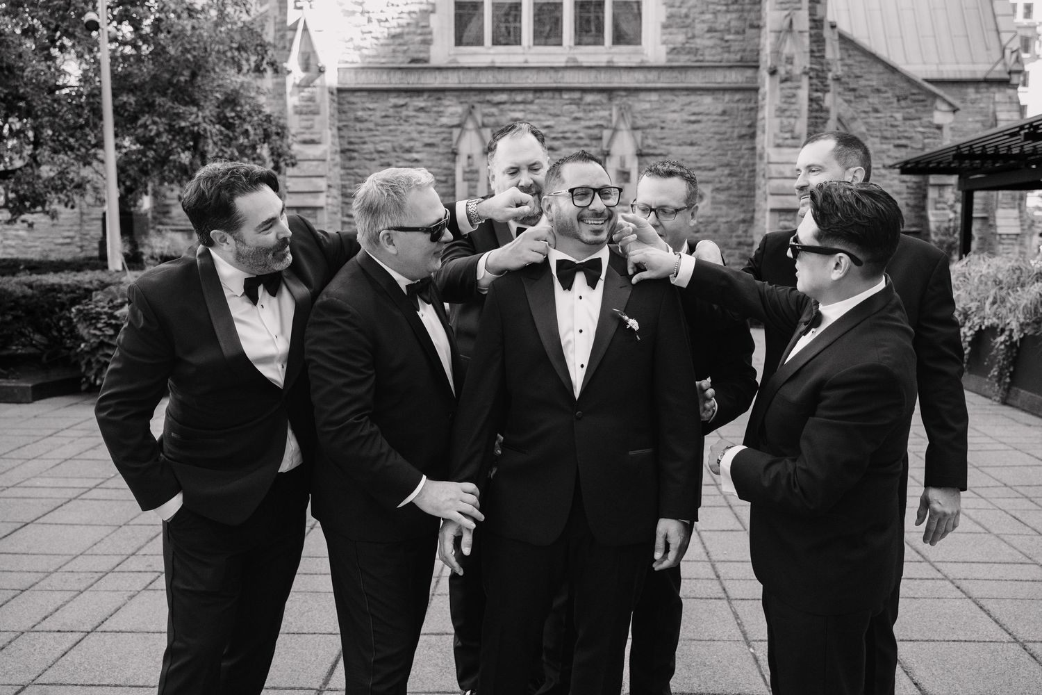 Groom surrounded by smiling groomsmen in black tuxedos outside stone church, black and white photo.