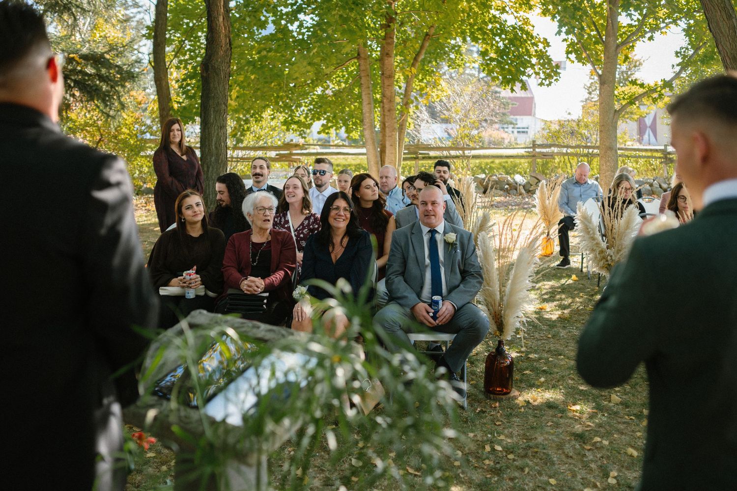 Wedding guests seated outdoors under trees watching the ceremony on a sunny autumn day.