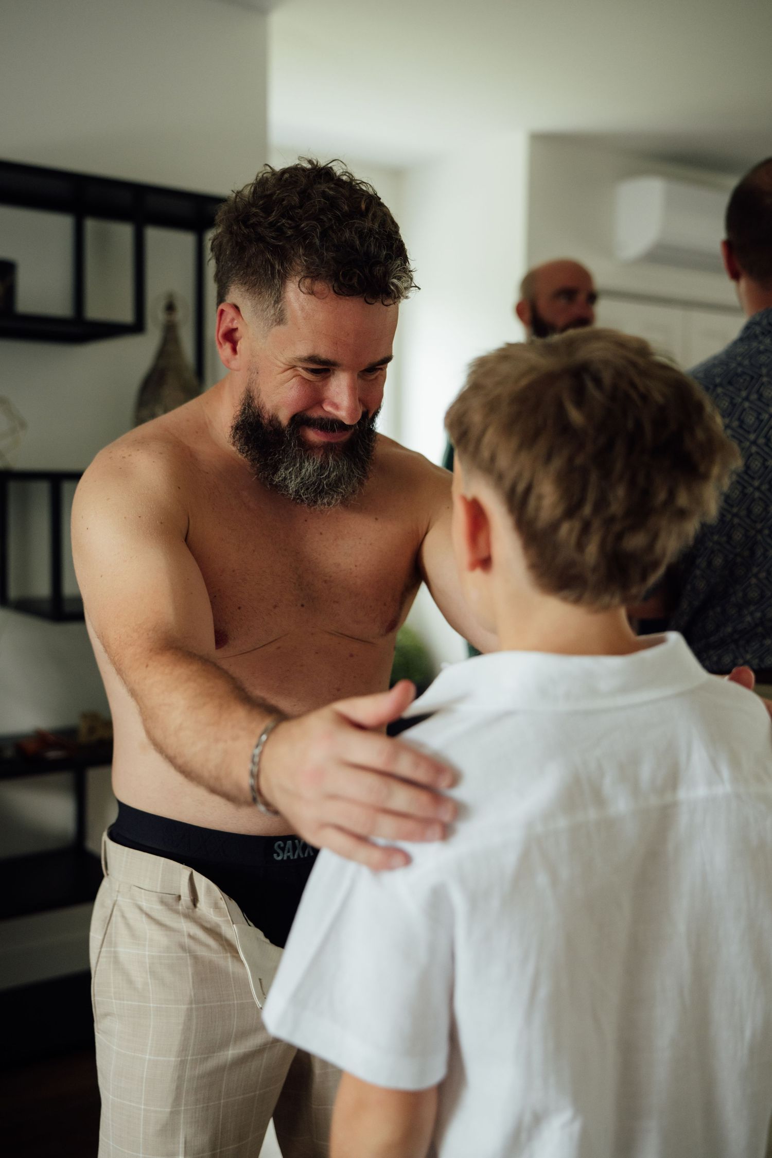 Shirtless bearded man placing hands on young boy's shoulders indoors during wedding preparations.