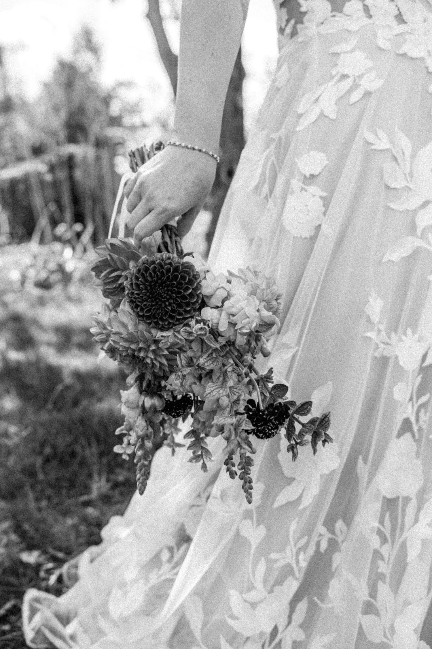 Black and white close-up of bride holding a dahlia and wildflower bouquet in a floral lace gown.