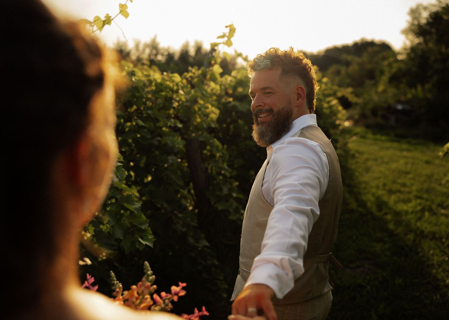 A bearded groom in a tan vest smiles warmly while holding hands in golden sunset garden light.