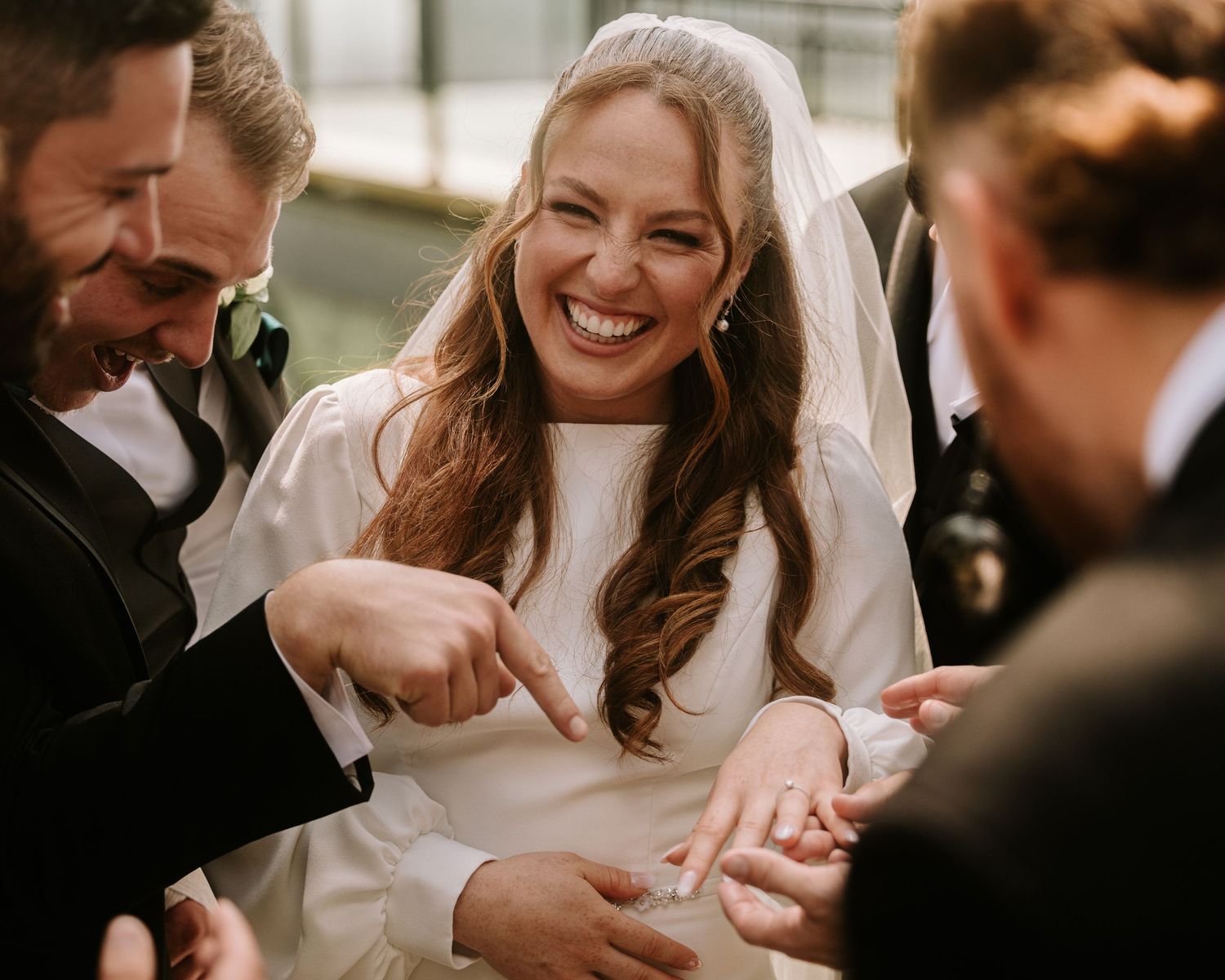 Bride in white gown and veil laughs joyfully with groomsmen pointing at her wedding ring during celebration.