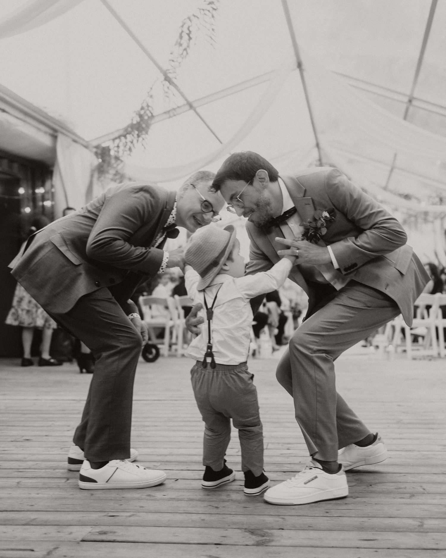 Two groomsmen in suits lean down to interact with a toddler in suspenders on a wooden dance floor at a wedding.