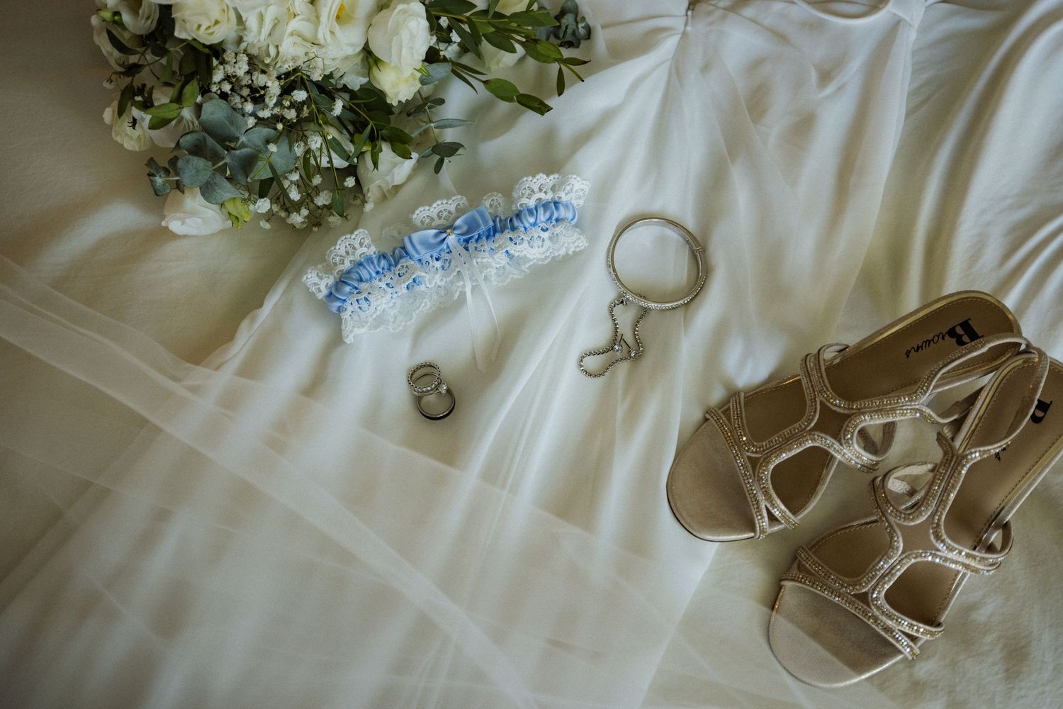 Bridal flat lay with white garter, sandals, rings, and floral bouquet on wedding dress veil.