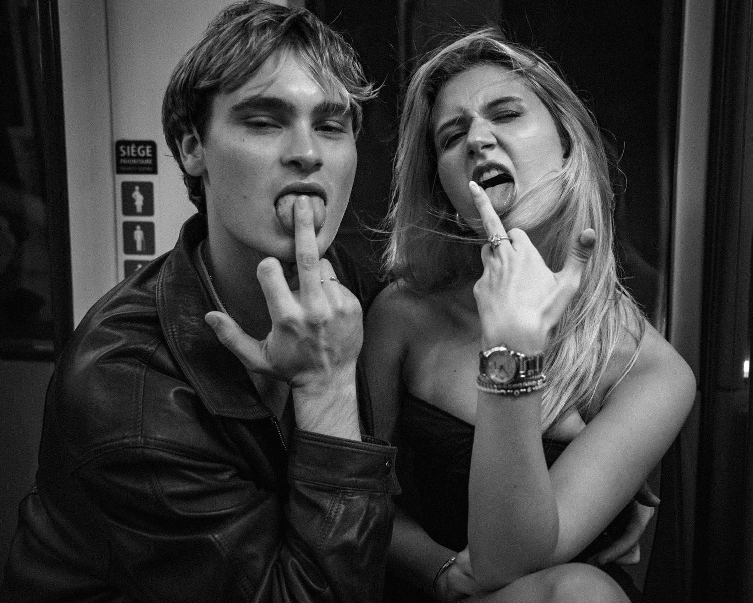 Black and white photo of a young couple playfully sticking out their tongues on a subway train.