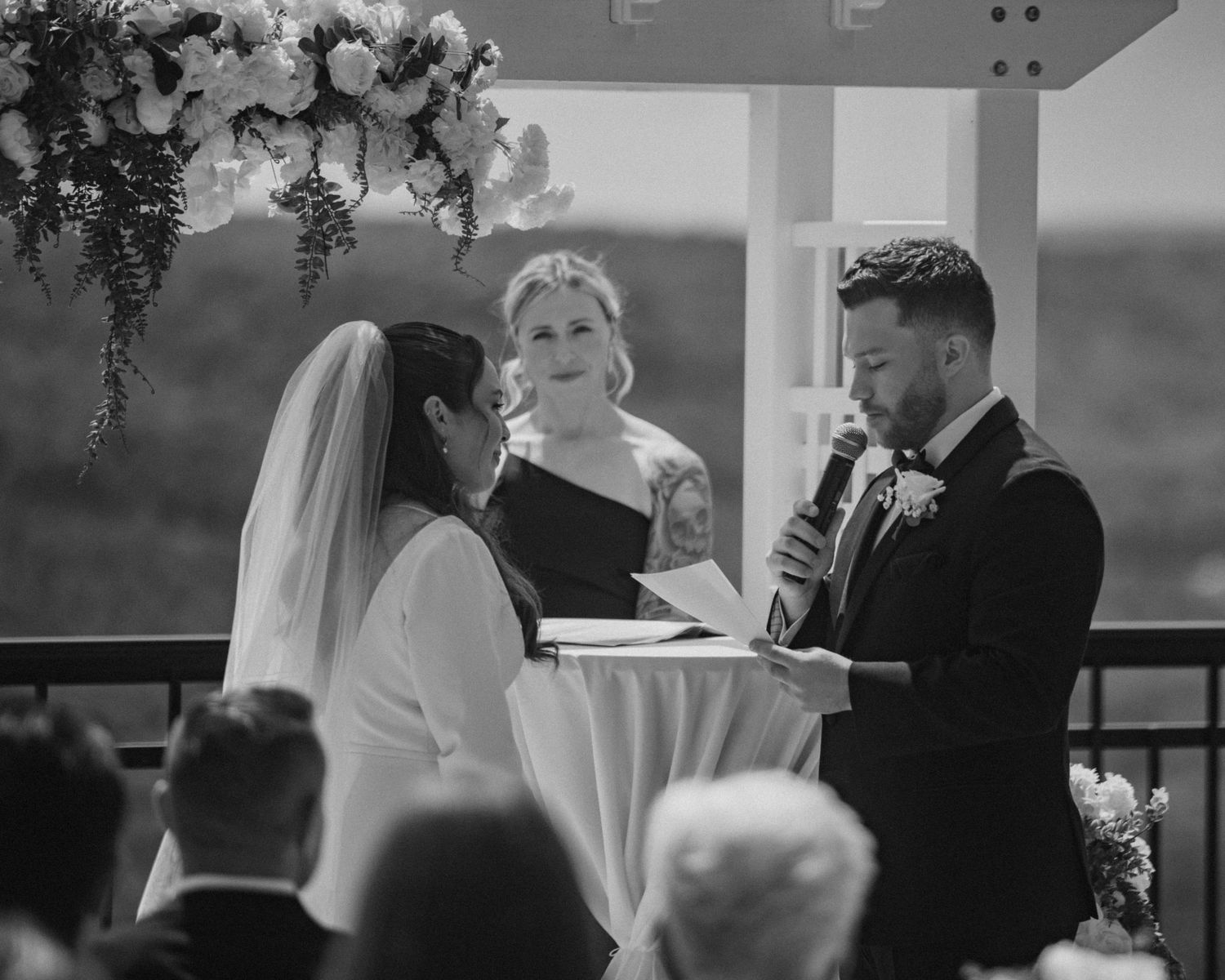 Black and white photo of groom reading vows to bride at outdoor wedding ceremony with officiant and floral arch backdrop.